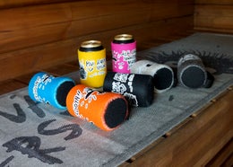 Refroidisseurs de canette Le H&eacute;risson Juron de diff&eacute;rentes couleurs sur une serviette pour banc de sauna Hyv&auml; perse.